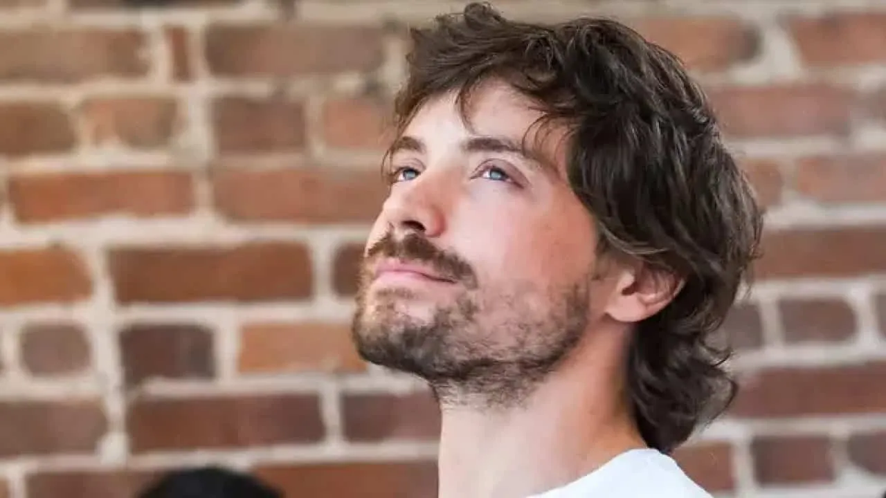 Side profile of a man with medium-length brown hair and a beard against a brick wall background